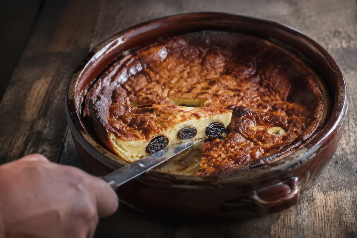 Far breton aux pruneaux dans un plat en terre vernissée, une part coupée qui laisse voir les pruneaux, couteau et main vue de dos, table en bois de ferme, lumière du jour