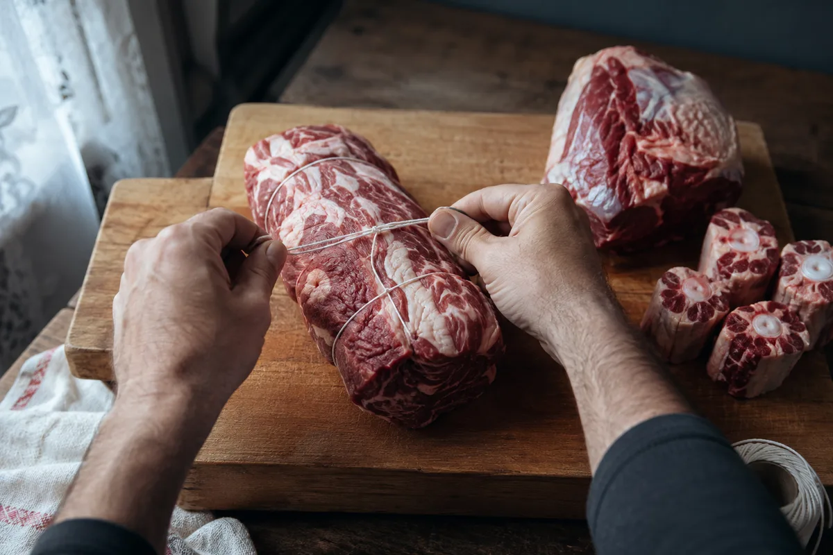 Mains vues de dos qui ficellent un morceau de plat de côte cru sur une planche en bois, gîte et tronçons de queue à côté, lumière de jour