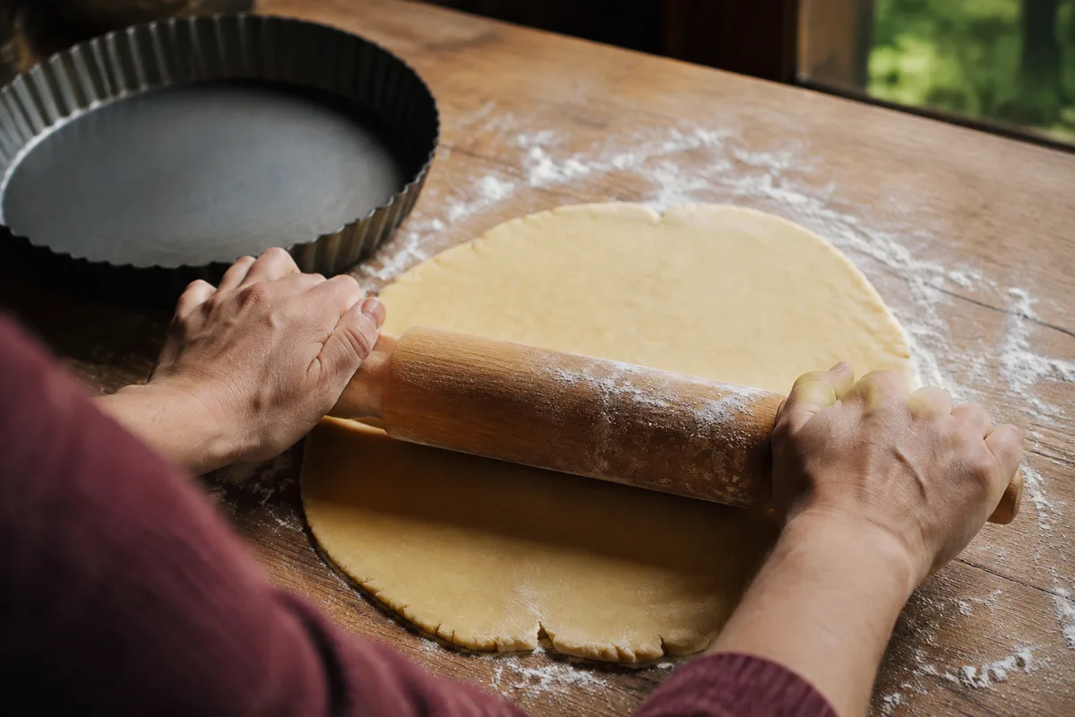 Mains vues de dos qui étalent une pâte brisée au rouleau sur un plan de travail fariné, moule à tarte en tôle vide à côté, lumière de fenêtre