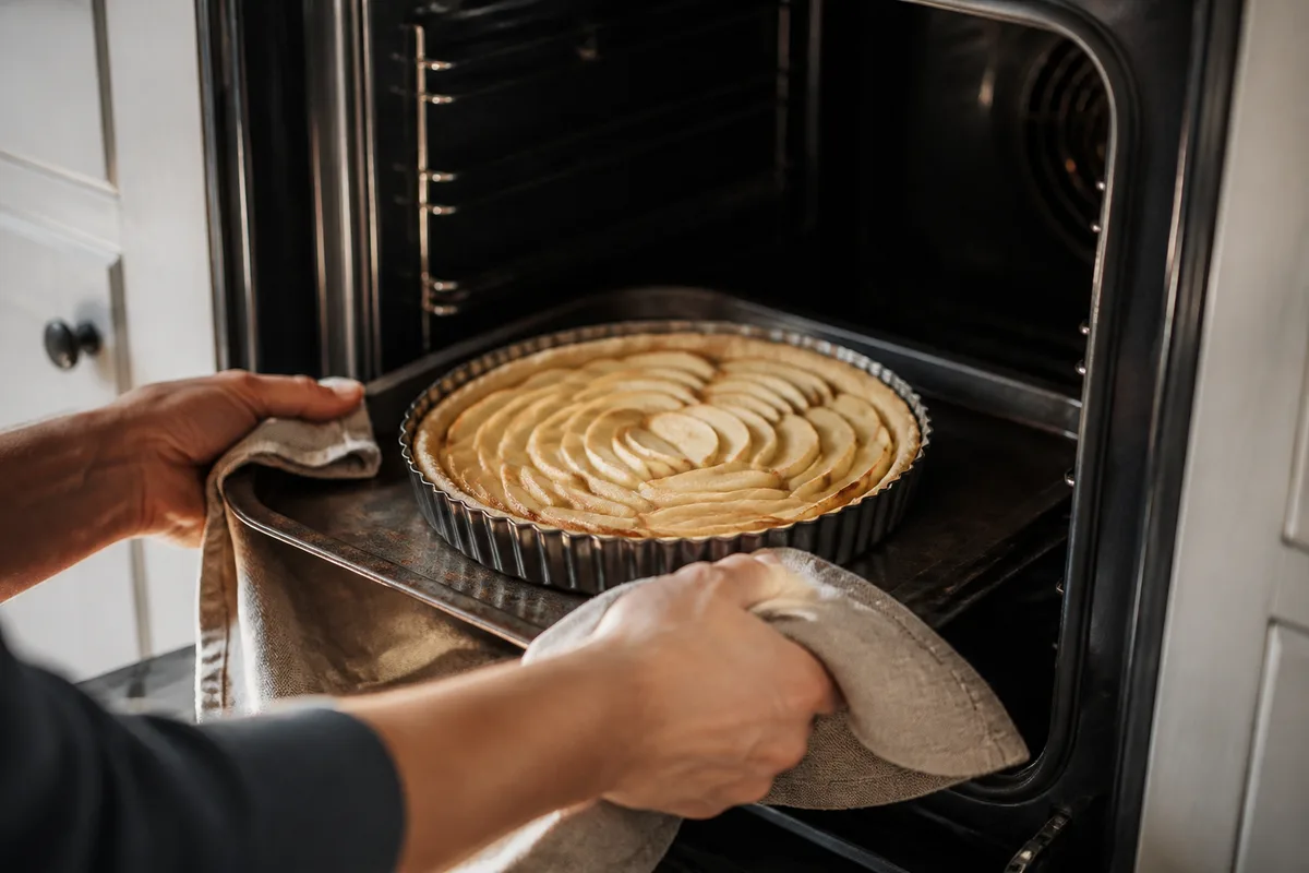 Tarte aux pommes en lamelles enfournée sur une plaque en métal tout en bas d'un four de maison éteint, porte ouverte, mains vues de dos qui tiennent le moule avec un torchon